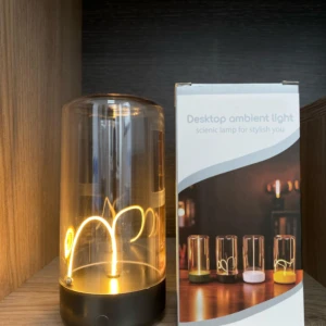 Lámpara táctil imantada de sobremesa con cuerpo de vidrio cilíndrico y filamento luminoso curvo, junto a su caja de empaque con diseño decorativo.