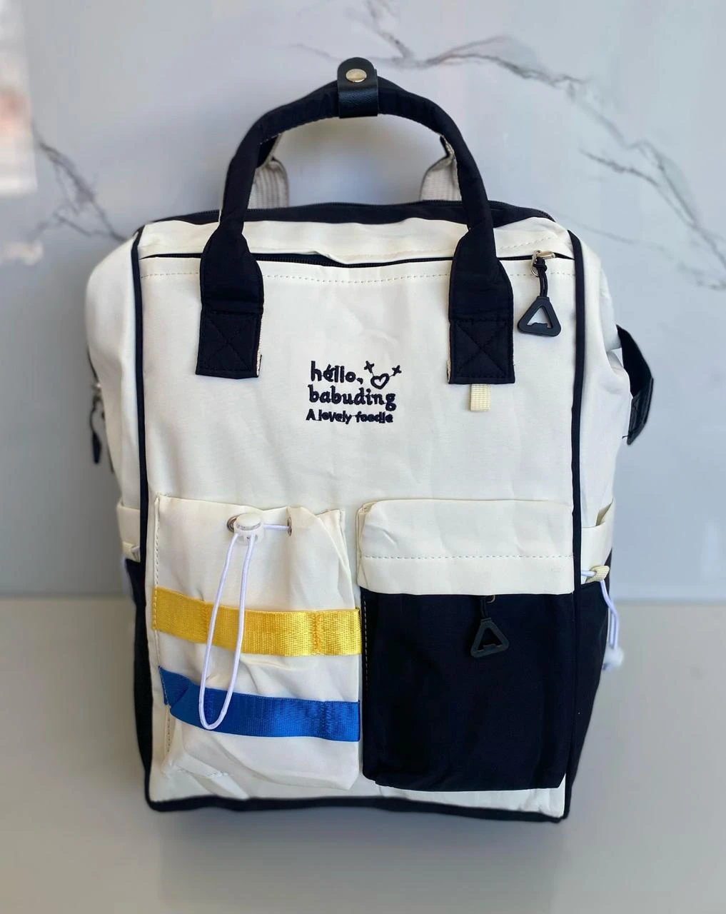 Pañalera blanca con negro, dos bolsillos frontales, uno con rayas amarillas y azules.