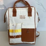 Pañalera blanca y café con bordes marrones, dos bolsillos frontales, asas de cuero y bordado "hello, babuding".