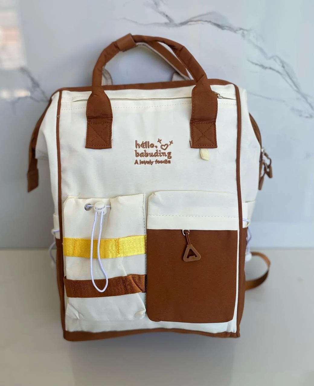 Pañalera blanca y café con bordes marrones, dos bolsillos frontales, asas de cuero y bordado "hello, babuding".