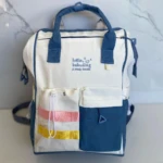 Pañalera color marfil y azul marino, con dos bolsillos frontales, uno con cintas de colores.