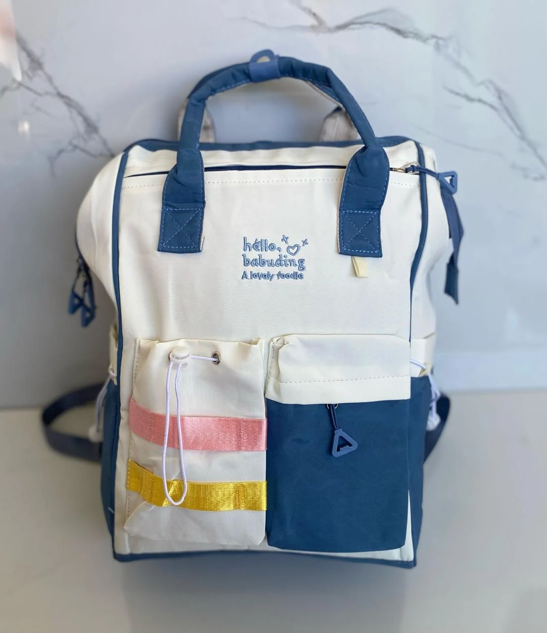 Pañalera color marfil y azul marino, con dos bolsillos frontales, uno con cintas de colores.