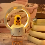 Lámpara LED recargable con aro de luz y figura de oso amarillo, base blanca sobre soporte metálico cobre, junto a contenedores apilados.