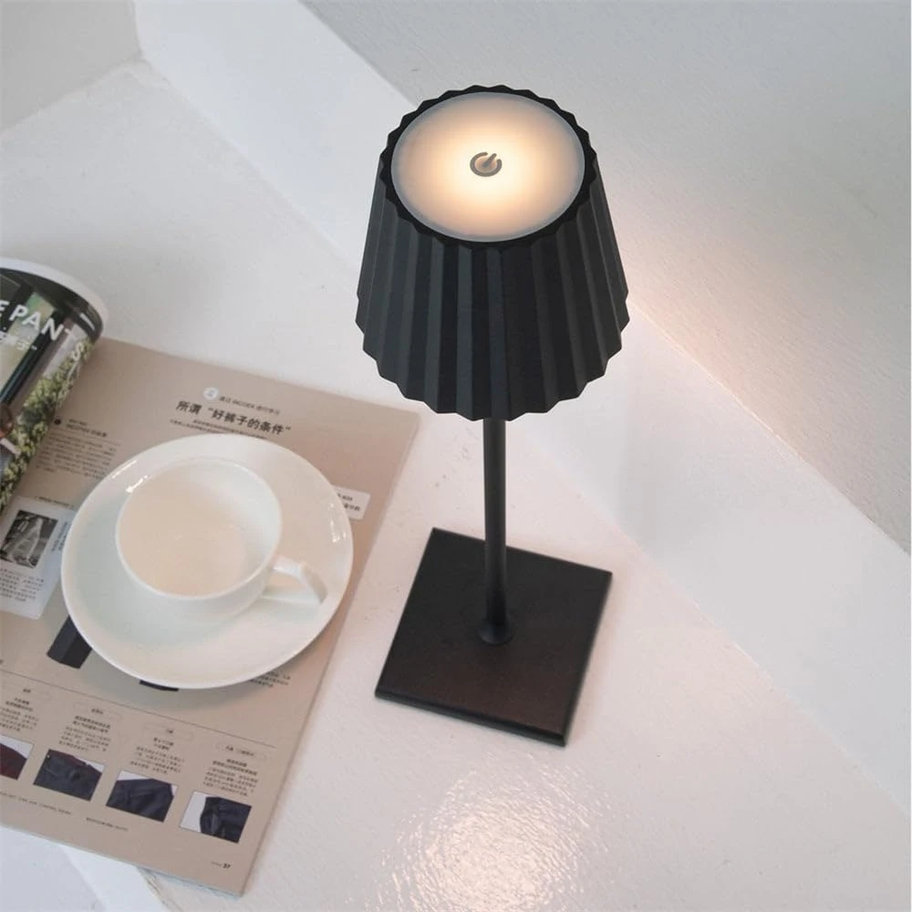 Lámpara LED inalámbrica de mesa color negro mate, con pantalla estriada, base cuadrada y botón táctil superior, ubicada junto a una taza y una revista.