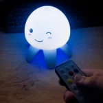 Lámpara de noche tipo medusa con luz LED azul y cara expresiva, controlada por un mando a distancia en primer plano sobre un escritorio de madera.