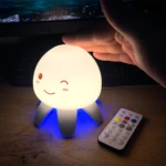 Lámpara de medusa F8 con luz blanca cálida y base azul, diseño de cara sonriente, operada con control remoto inalámbrico sobre una superficie de madera.