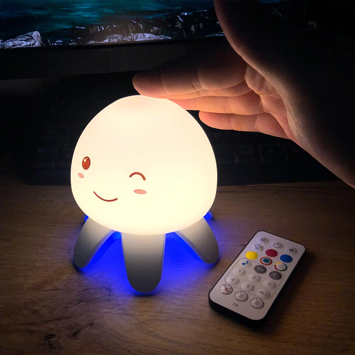 Lámpara de medusa F8 con luz blanca cálida y base azul, diseño de cara sonriente, operada con control remoto inalámbrico sobre una superficie de madera.