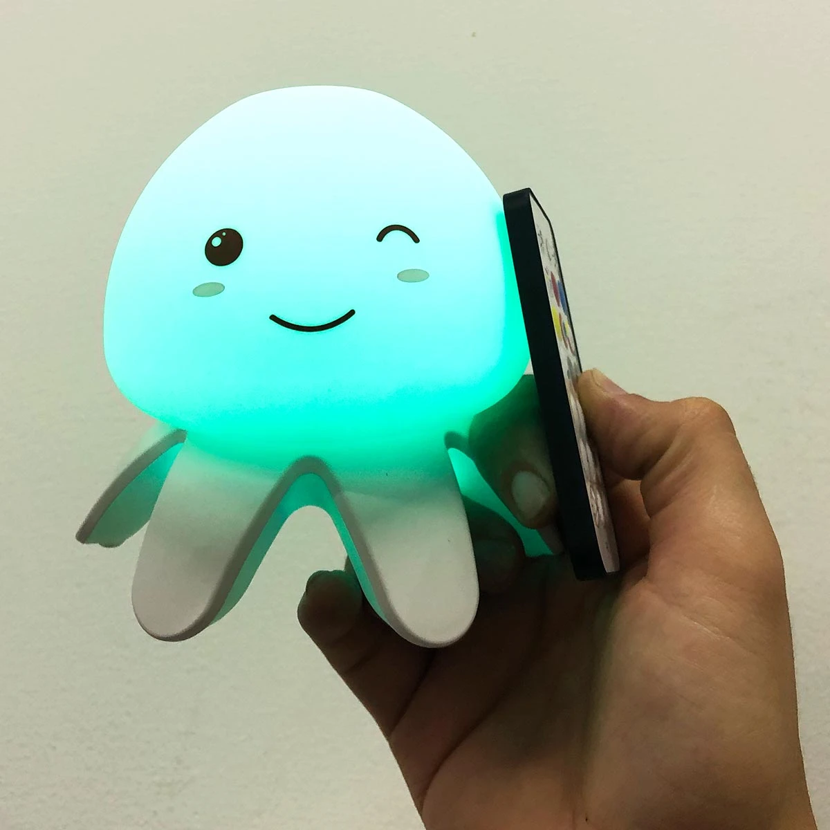 Lámpara de noche LED con forma de medusa color aguamarina, sostiene un control remoto pequeño en una mano humana, acabado en silicona suave y mate.