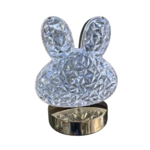 Lámpara LED de mesa con figura de cabeza de conejo en acrílico facetado cristalino sobre base metálica dorada reflectiva.