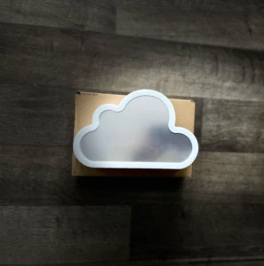 Lámpara decorativa con forma de nube blanca y panel difusor central, ubicada sobre su caja de cartón original, vista desde un ángulo superior.