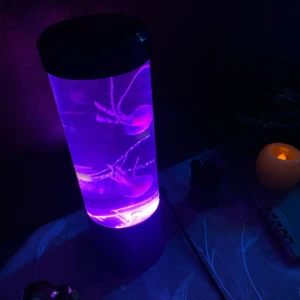 Lámpara LED cilíndrica con dos medusas artificiales flotando, iluminada en tonos violeta, ubicada sobre una mesa junto a una vela decorativa encendida.