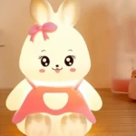 Lámpara infantil LED de silicona con forma de conejito blanco, ojos grandes, lazo rosa y vestido naranja, ideal como luz de noche para niños.