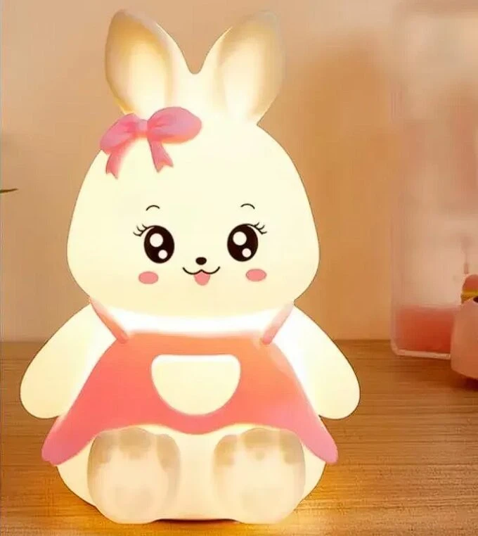 Lámpara infantil LED de silicona con forma de conejito blanco, ojos grandes, lazo rosa y vestido naranja, ideal como luz de noche para niños.
