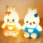 Par de lámparas LED infantiles en forma de conejos de silicona. Izquierda con vestido naranja y moño, derecha con overol azul y gorro, sobre mesa.