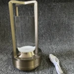 Lámpara de luz táctil metálica color bronce con cilindro de vidrio transparente y cable de carga blanco, sobre superficie de textura gris.