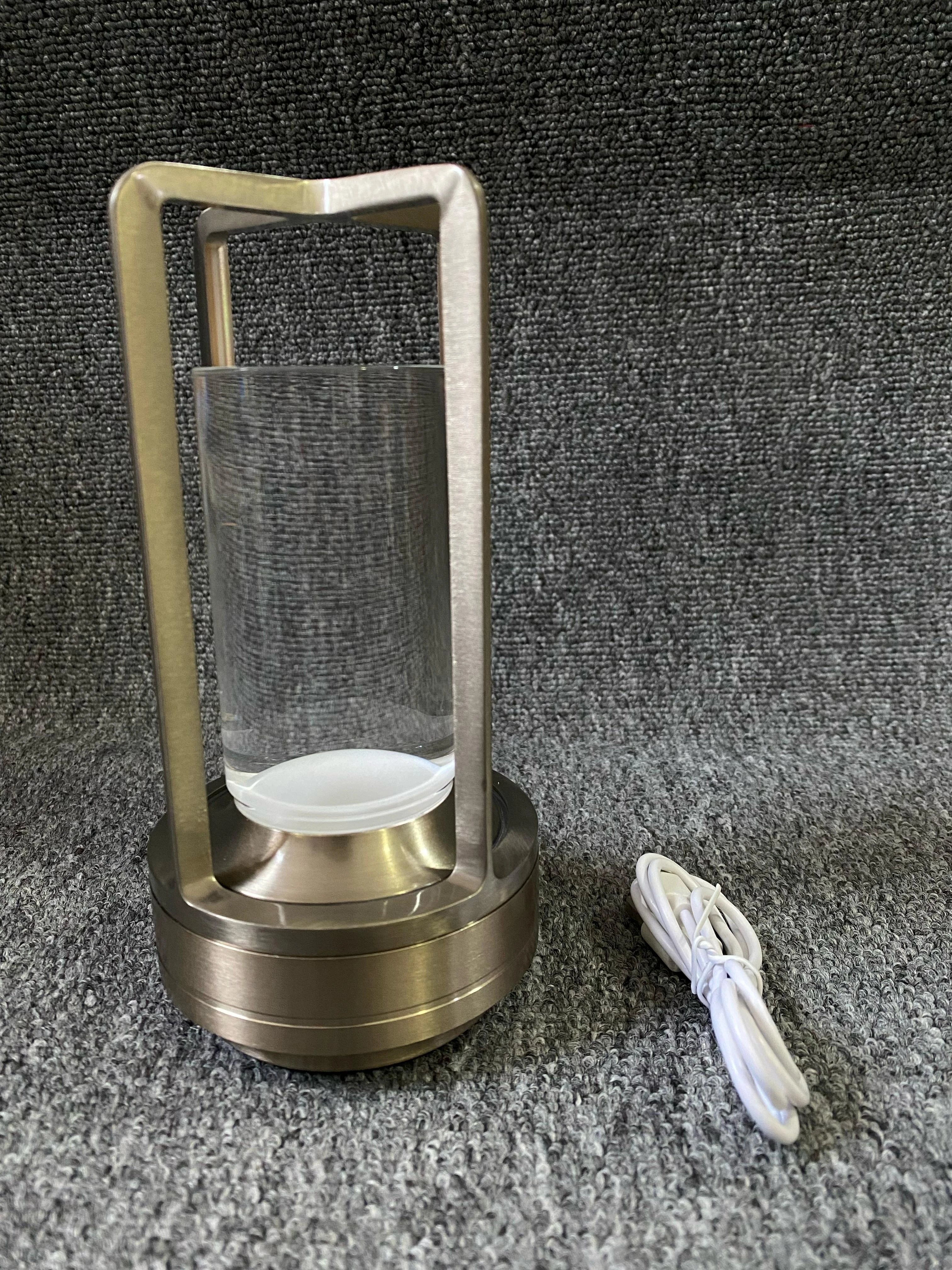 Lámpara de luz táctil metálica color bronce con cilindro de vidrio transparente y cable de carga blanco, sobre superficie de textura gris.