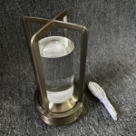 Lámpara de luz táctil con estructura metálica color bronce, difusor de vidrio cilíndrico transparente y cable de carga blanco incluido sobre fondo gris.