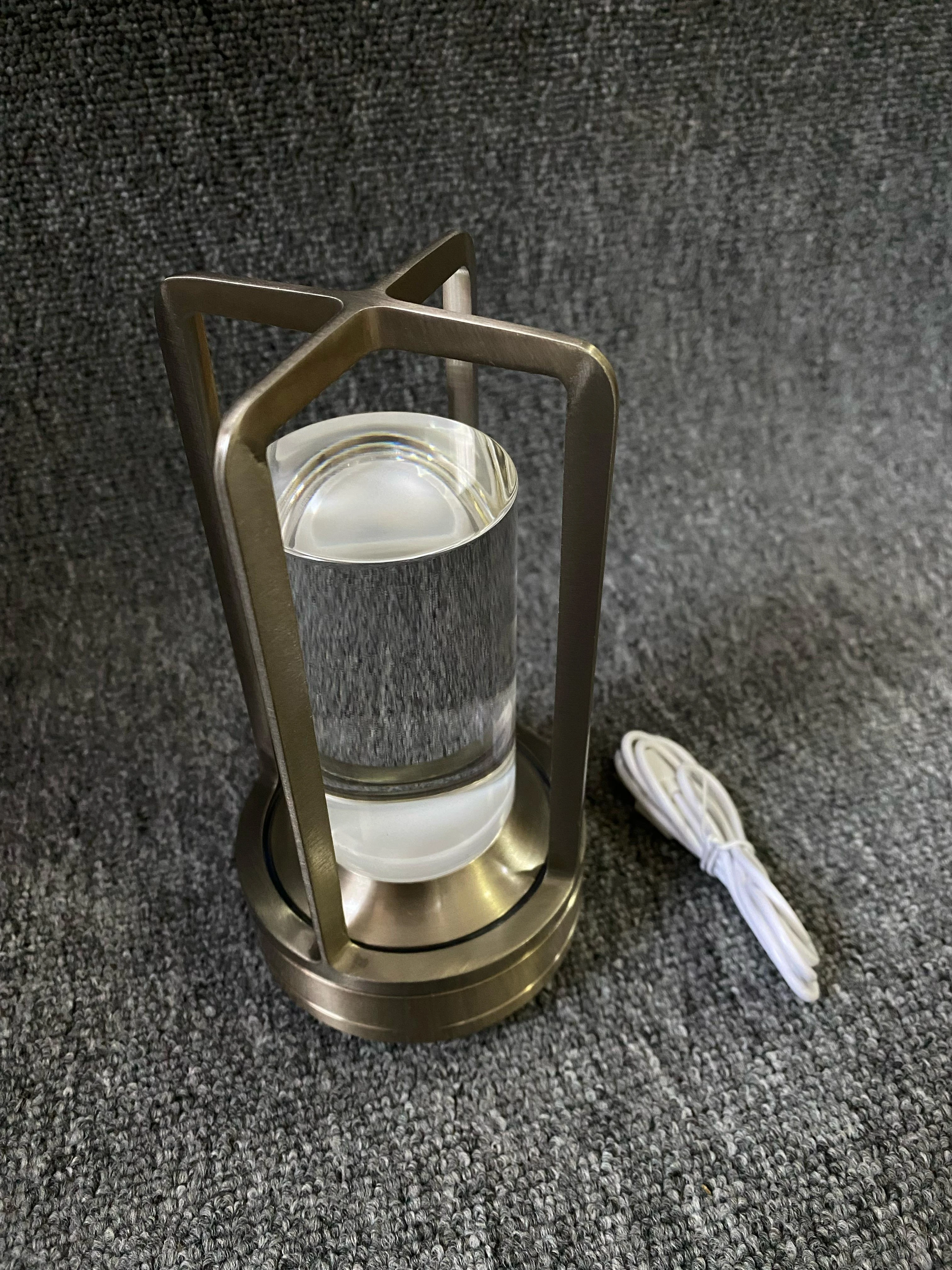 Lámpara de luz táctil con estructura metálica color bronce, difusor de vidrio cilíndrico transparente y cable de carga blanco incluido sobre fondo gris.