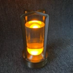 Lámpara de luz táctil con diseño cilíndrico de base metálica cepillada, estructura de marco en cruz y difusor transparente con iluminación LED cálida.