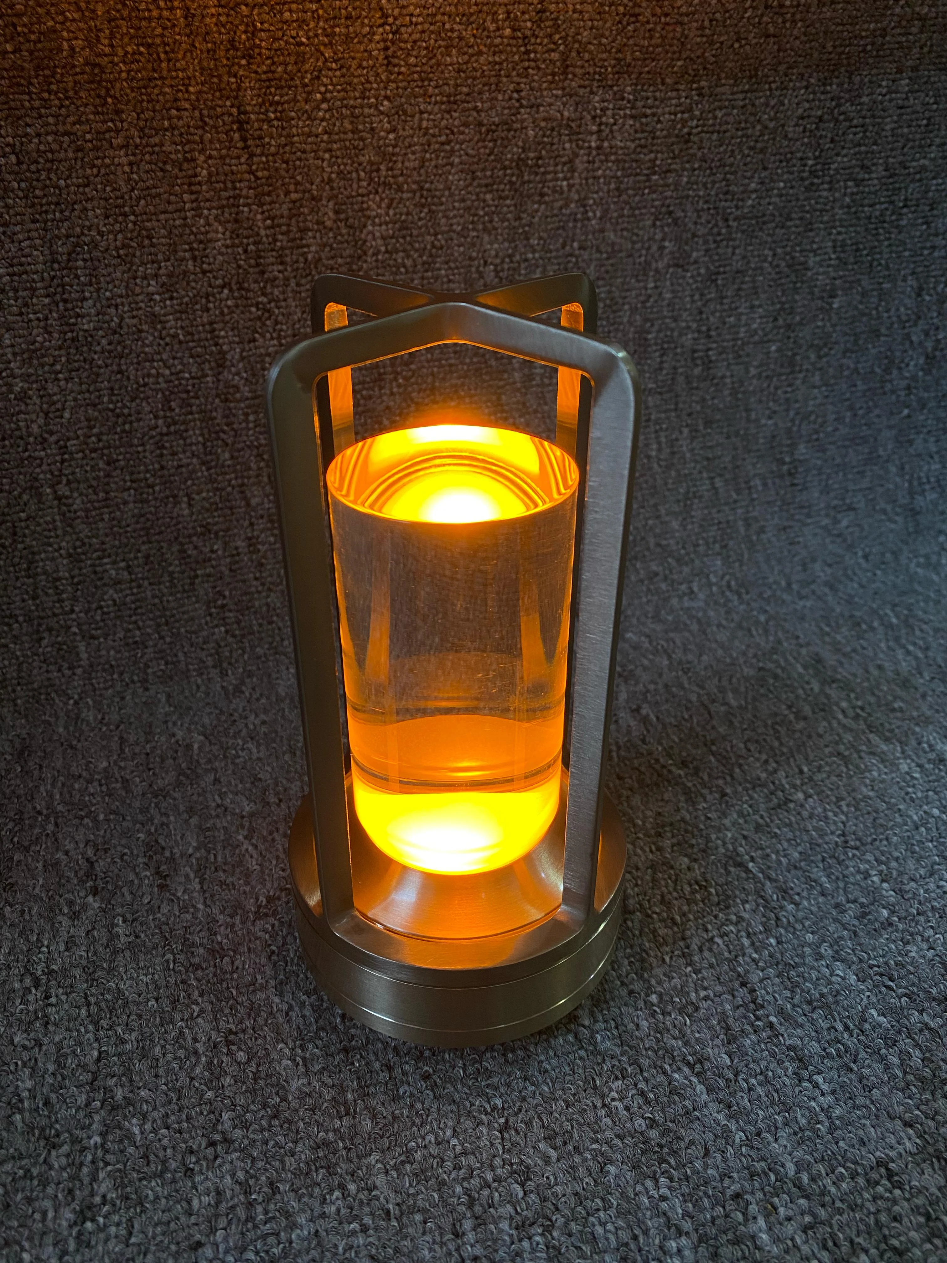 Lámpara de luz táctil con diseño cilíndrico de base metálica cepillada, estructura de marco en cruz y difusor transparente con iluminación LED cálida.