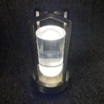 Lámpara de mesa táctil con base y estructura metálica color níquel, cuerpo cilíndrico de acrílico transparente y luz LED cálida encendida sobre fondo gris.