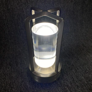 Lámpara de mesa táctil con base y estructura metálica color níquel, cuerpo cilíndrico de acrílico transparente y luz LED cálida encendida sobre fondo gris.