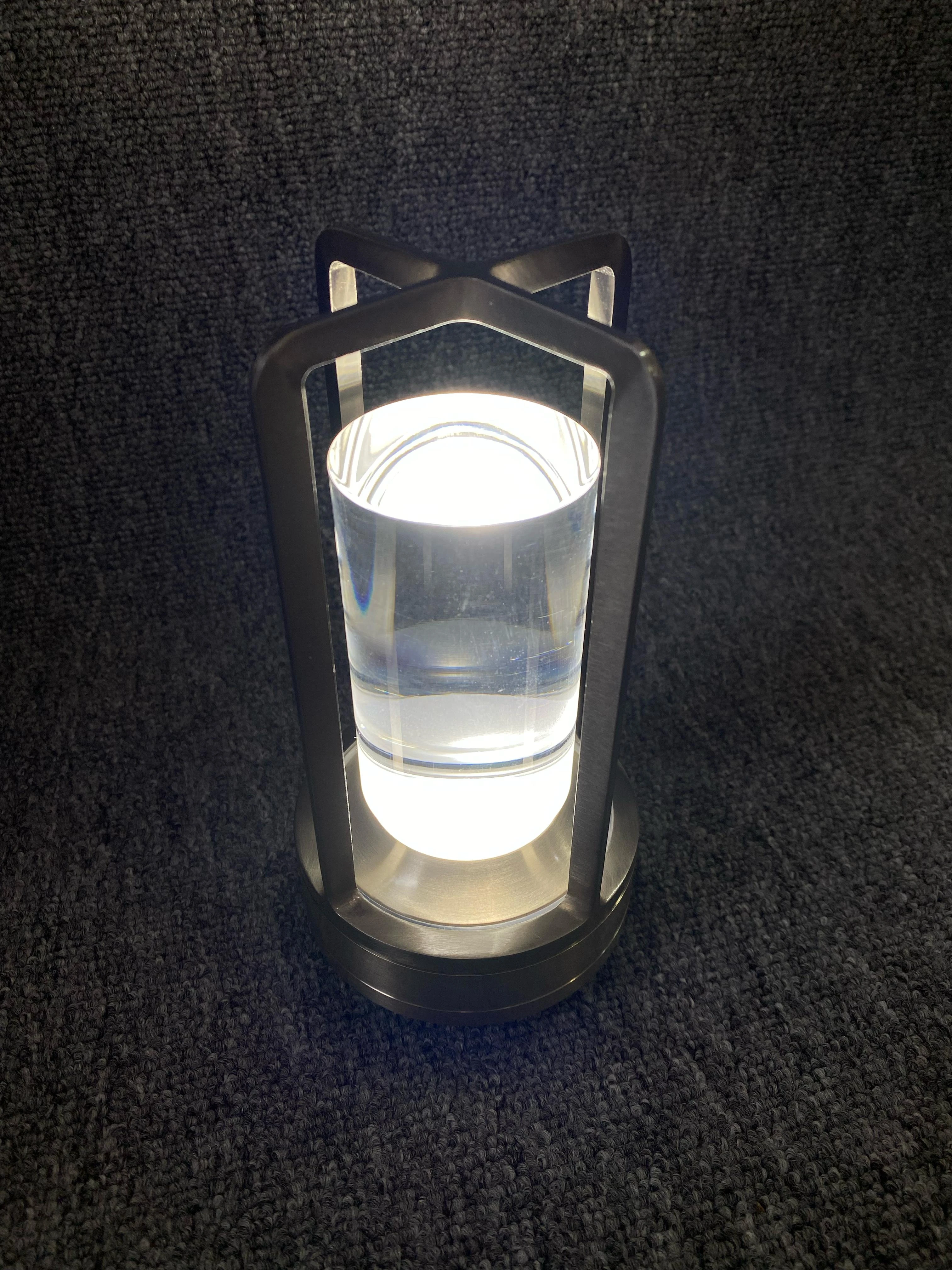 Lámpara de mesa táctil con base y estructura metálica color níquel, cuerpo cilíndrico de acrílico transparente y luz LED cálida encendida sobre fondo gris.