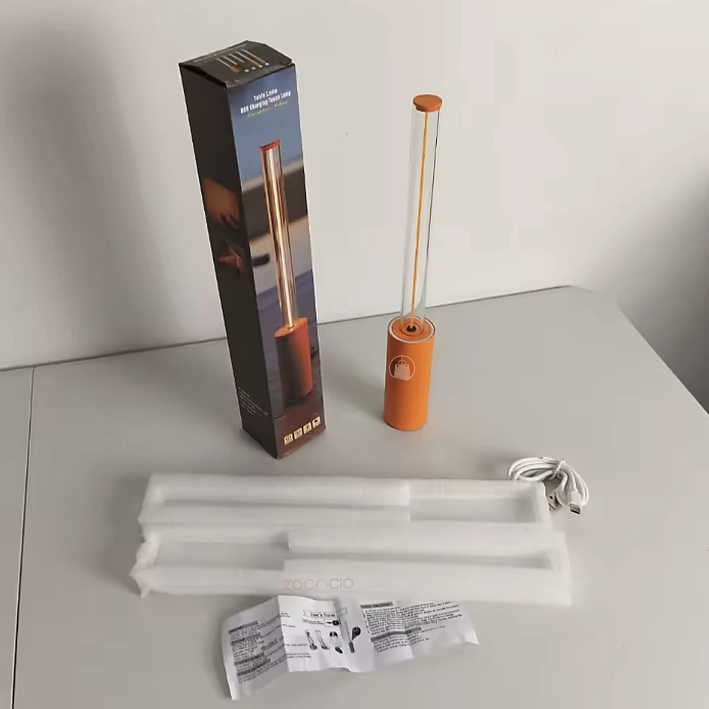 Mini lámpara de ambiente minimalista color naranja, incluye caja, cable de carga USB, empaque protector y manual de usuario sobre una mesa blanca.