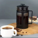 Cafetera prensa francesa de vidrio y metal negro 600ml, con taza de café y trozo de pastel.