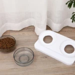 Comedero doble elevado blanco para mascota, con dos cuencos transparentes: uno con croquetas y otro vacío, sobre piso de madera.