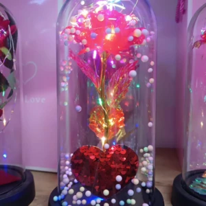 Lámpara de rosa roja brillante con luces LED y base de corazón de lentejuelas, decorada con bolitas multicolores bajo cúpula de cristal.