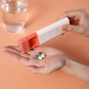 Organizador de pastillas de viaje blanco y naranja con 3 espacios, mostrando cápsulas en la mano y vaso de agua.