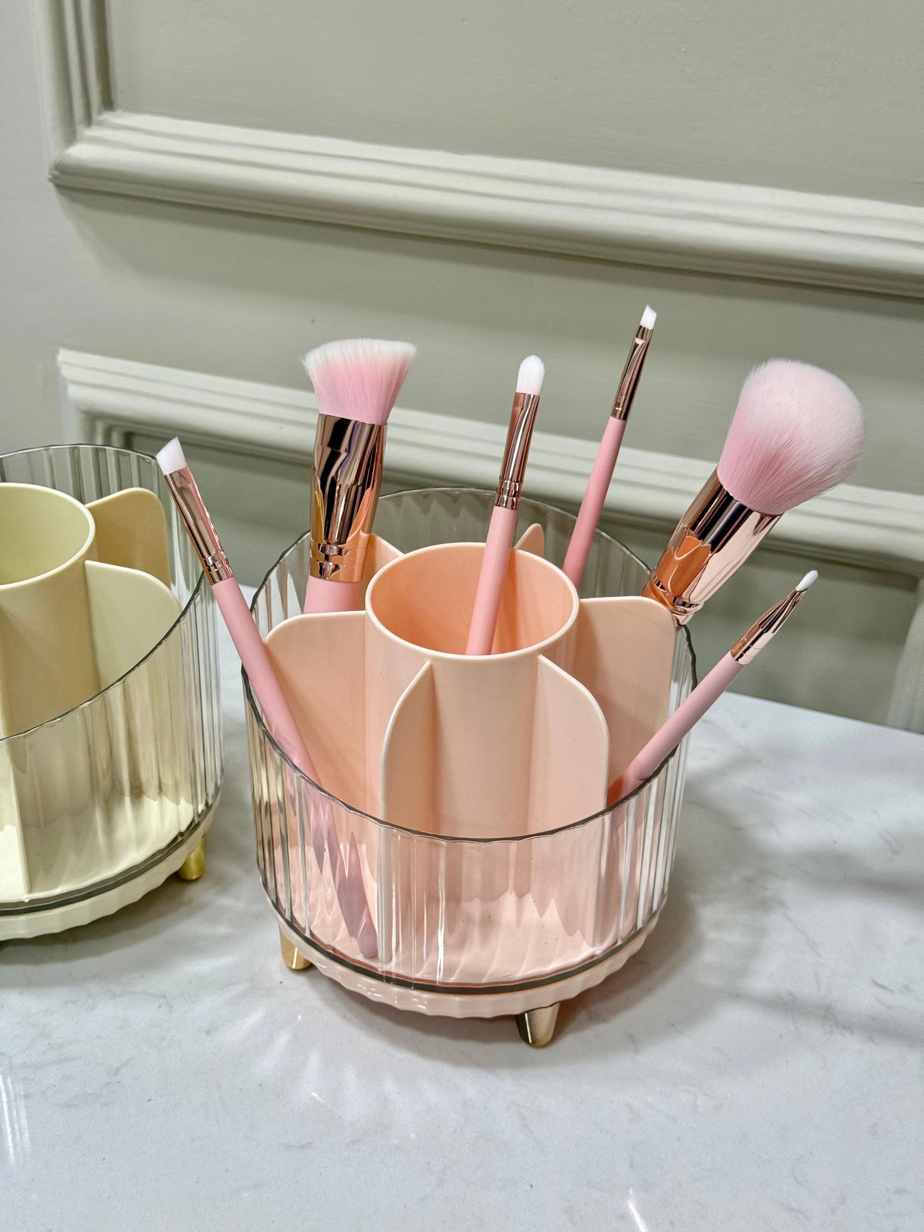 Organizador de cosméticos rosa con compartimentos y brochas de maquillaje, base transparente con patas doradas.