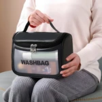 Cosmetiquera negra tipo washbag, transparente, con texto "WASHBAG" y cremallera plateada, sostenida por manos.