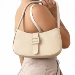 Bolso hombro estilo baguette beige claro, piel sintética brillante, con hebilla cuadrada decorativa frontal.