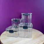Juego de 4 organizadores de cocina transparentes con tapas negras, sobre mesa rústica y fondo morado.