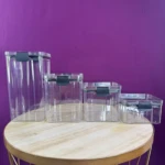 Cuatro organizadores de cocina rectangulares transparentes de plástico con tapa negra sobre mesa redonda dorada y fondo morado.
