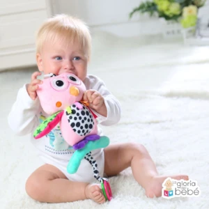 Bebé rubio con ojos azules muerde peluche de búho rosa con sonajero en pie.