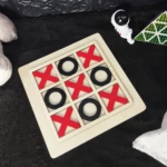 Juego de mesa Triqui pequeño de madera con X rojas y O negras en tablero de madera claro, sobre alfombra oscura.