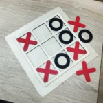 Tablero de madera para juego Triqui con fichas rojas en forma de X y negras en forma de O, con el juego a medio terminar.