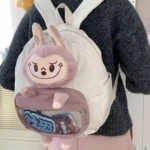 Morral blanco escolar con peluche de conejo malva en relieve y bolsillo frontal transparente con accesorios.