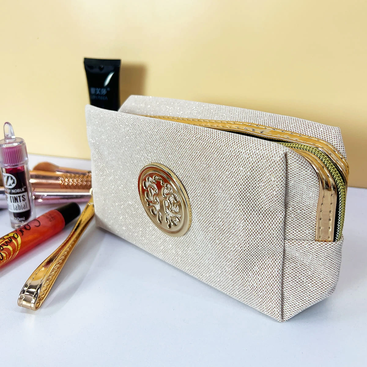 Cosmetiquera escarchada beige con detalle floral dorado, cremallera dorada, vista frontal.