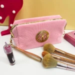 Cosmetiquera rosa texturizada con adorno dorado, correa, labial y brochas de maquillaje.