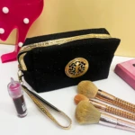 Cosmetiquera negra escarchada con logo dorado, asa de mano, labial y brochas de maquillaje en mesa blanca.