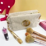 Cosmetiquera beige escarchada con asa dorada, adornos metálicos y brochas de maquillaje.