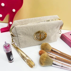Cosmetiquera beige escarchada con asa dorada, adornos metálicos y brochas de maquillaje.