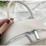 Morral bolso dama beige de cuero, con cremalleras plateadas y asa superior, mostrando el logo de @importacionesdac