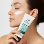 Mujer aplica crema protector solar facial Dr. Davey Collesen Herk Protect FPS 90+ en su mejilla, sosteniendo el tubo blanco de 100g.