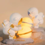 Lámpara de noche astronauta blanco con detalles dorados, cuidando una esfera luminosa con anillos dorados sobre base lunar.