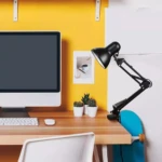 Lámpara de mesa negra articulada sujeta al borde de un escritorio de madera junto a un monitor y cactus.